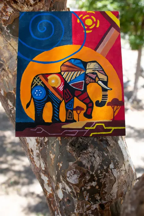 A painting of an elephant patterned in many colours against an orange sun, hung on a tree
