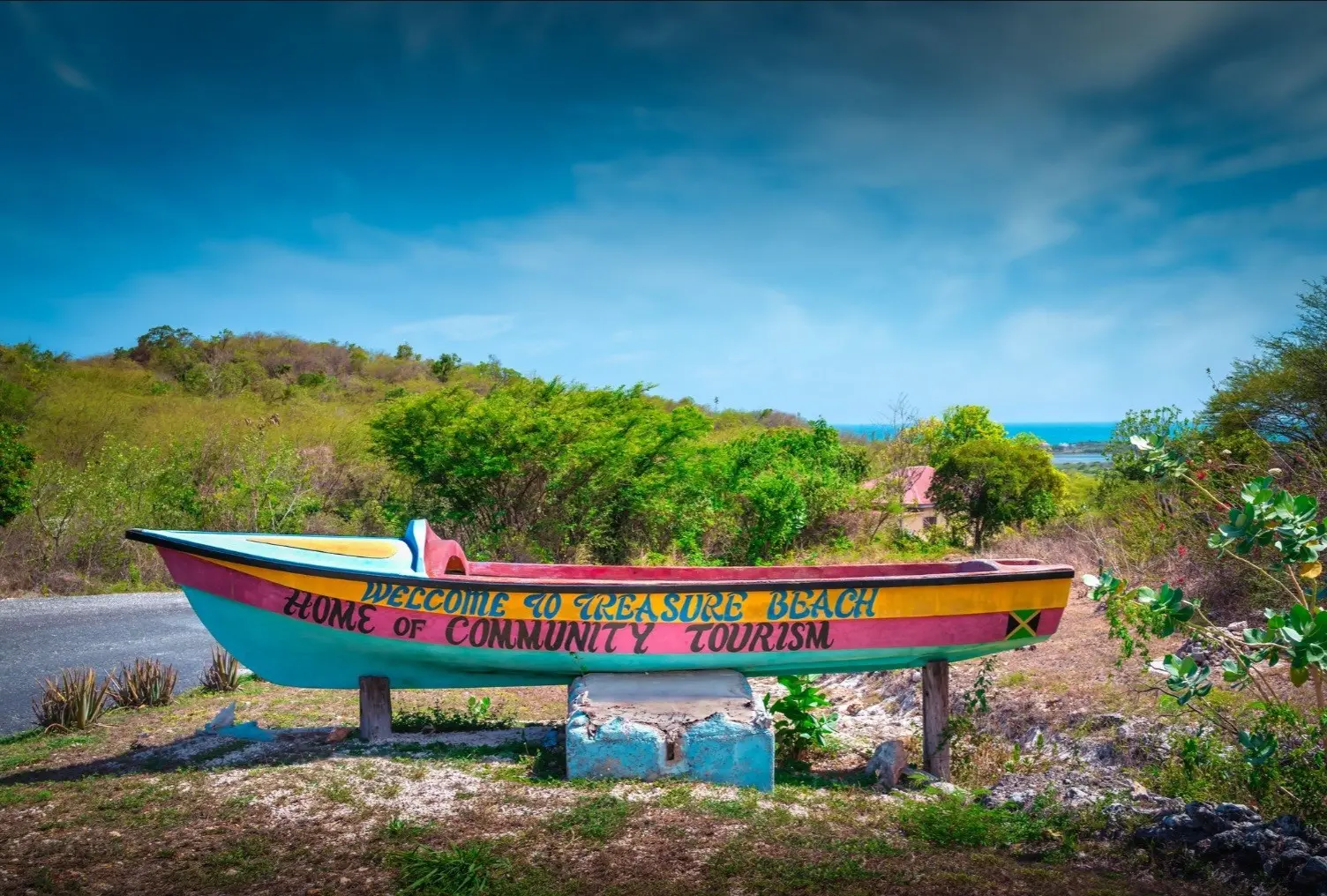 A painted fishing boat by the roadside reading “Welcome to Treasure Beach, Home of Community Tourism”, with the sea beyond