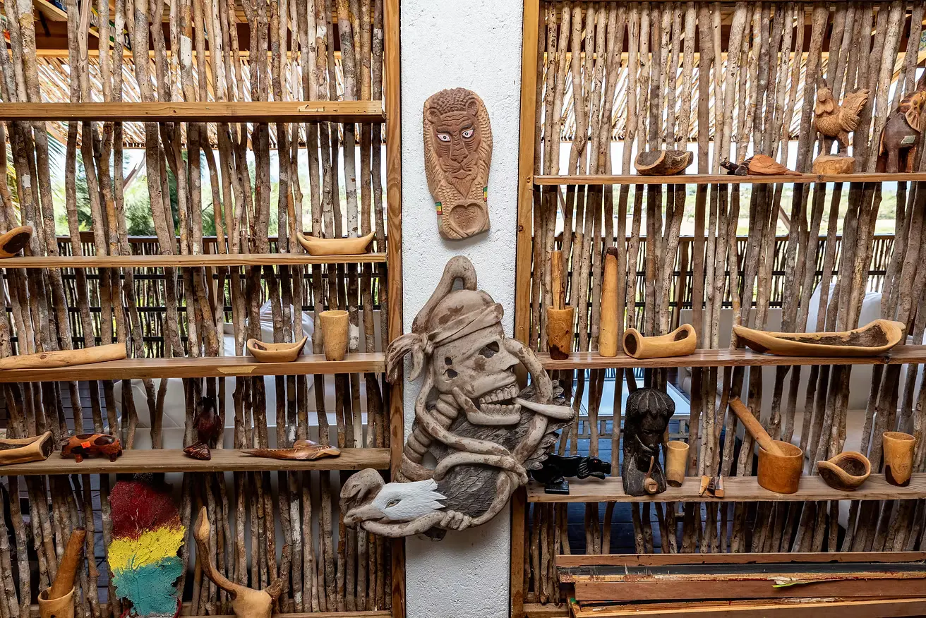 Open shelves of hand-carved wooden bowls, cups and figures against a woven stick wall, with a carved mask hanging on the white pillar