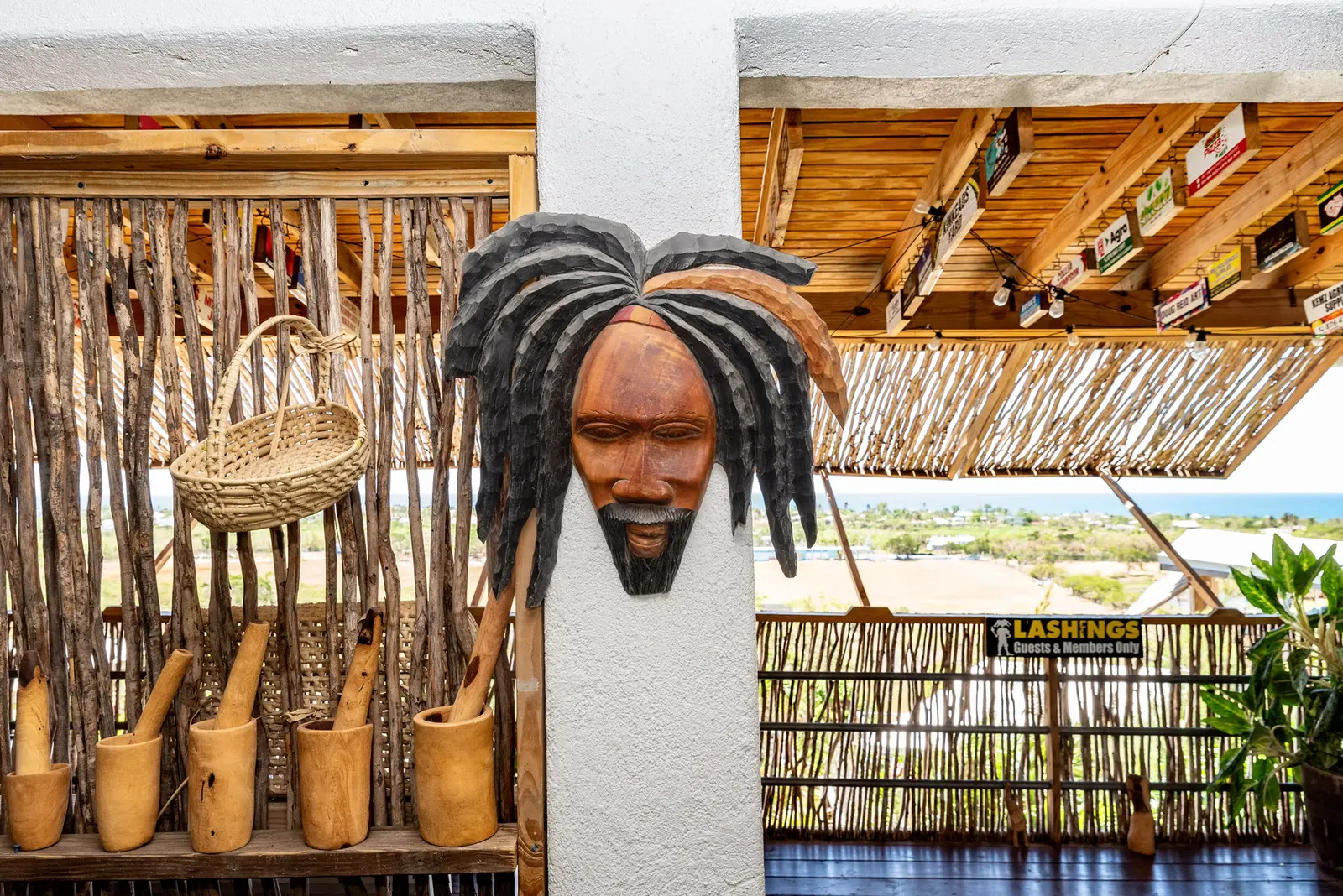 A carved wooden head with dreadlocks mounted on a post beside a basket and wooden mortars, with the sea and coast visible from the open balcony beyond