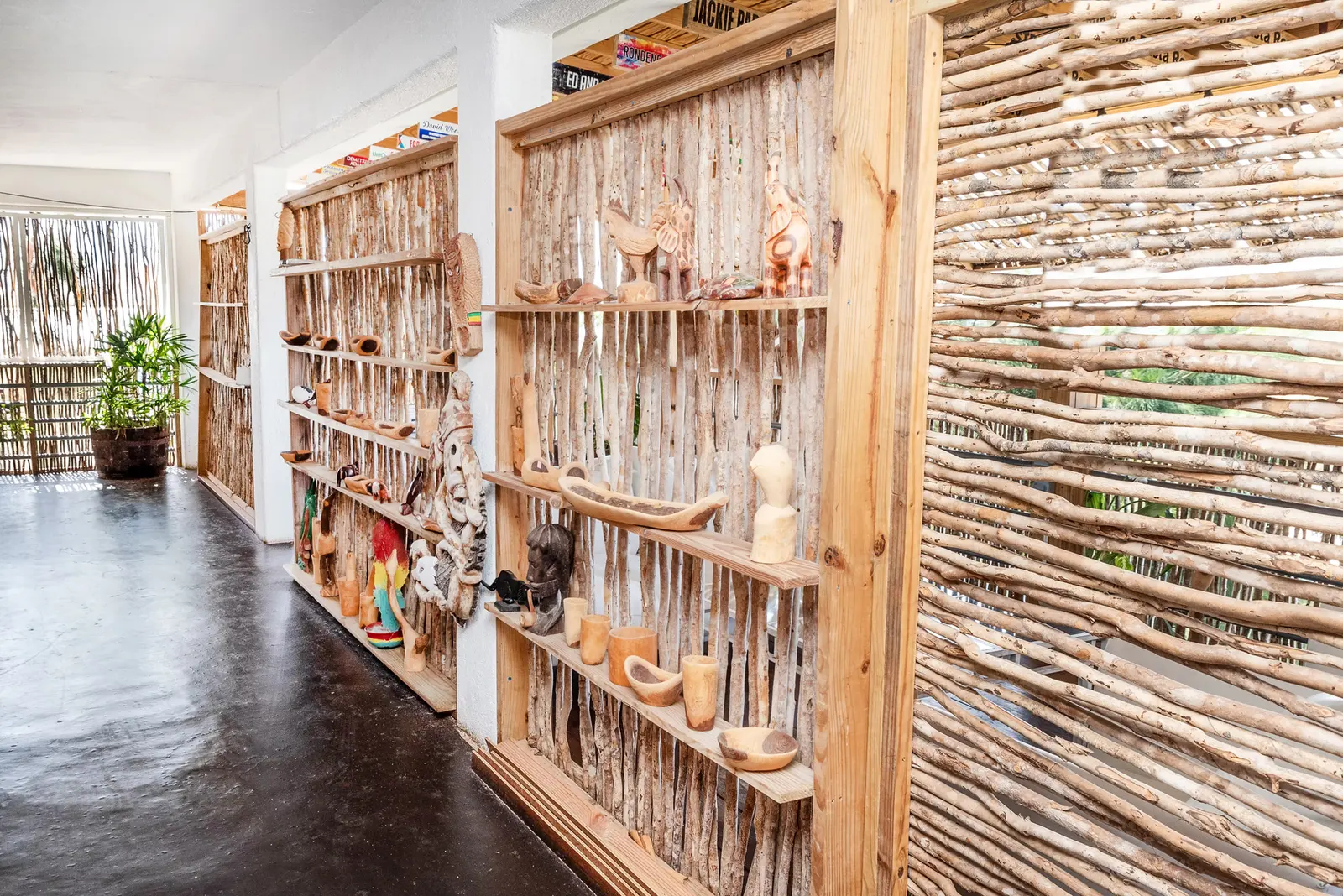 A passage lined with shelves of wood carvings along stick walls, with potted plants and a polished dark floor