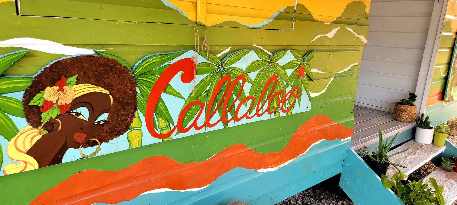 The painted wall beside the porch: a woman with an afro and a flower in her hair, the name Callaloo in red script among palm leaves, and waves of orange and teal
