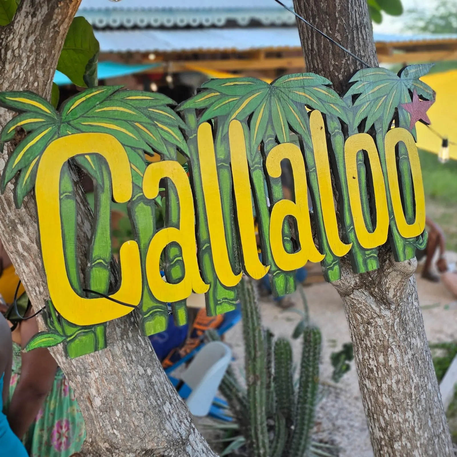 The yellow Callaloo lettering among green bamboo and palm leaves, hung between two tree trunks, with the shop's roof behind