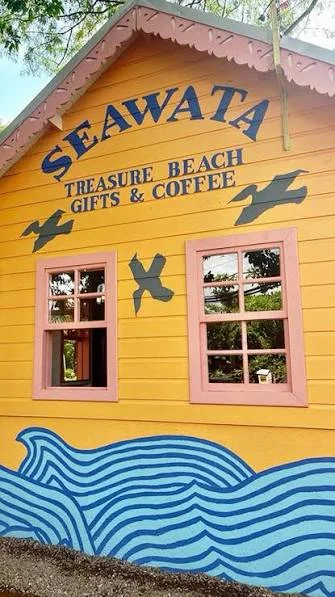The Seawata sign on a yellow wooden building with pink-trimmed windows: Treasure Beach Gifts and Coffee, with painted birds and a blue wave below