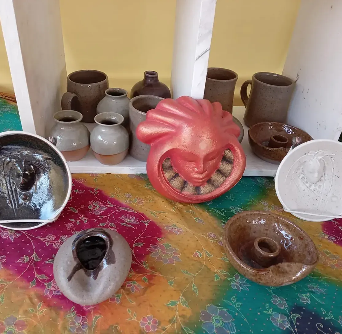 Handmade pottery on a white shelf and a bright cloth: brown and grey mugs and small vases, candle holders, round plates with moulded faces, and a pink glazed face