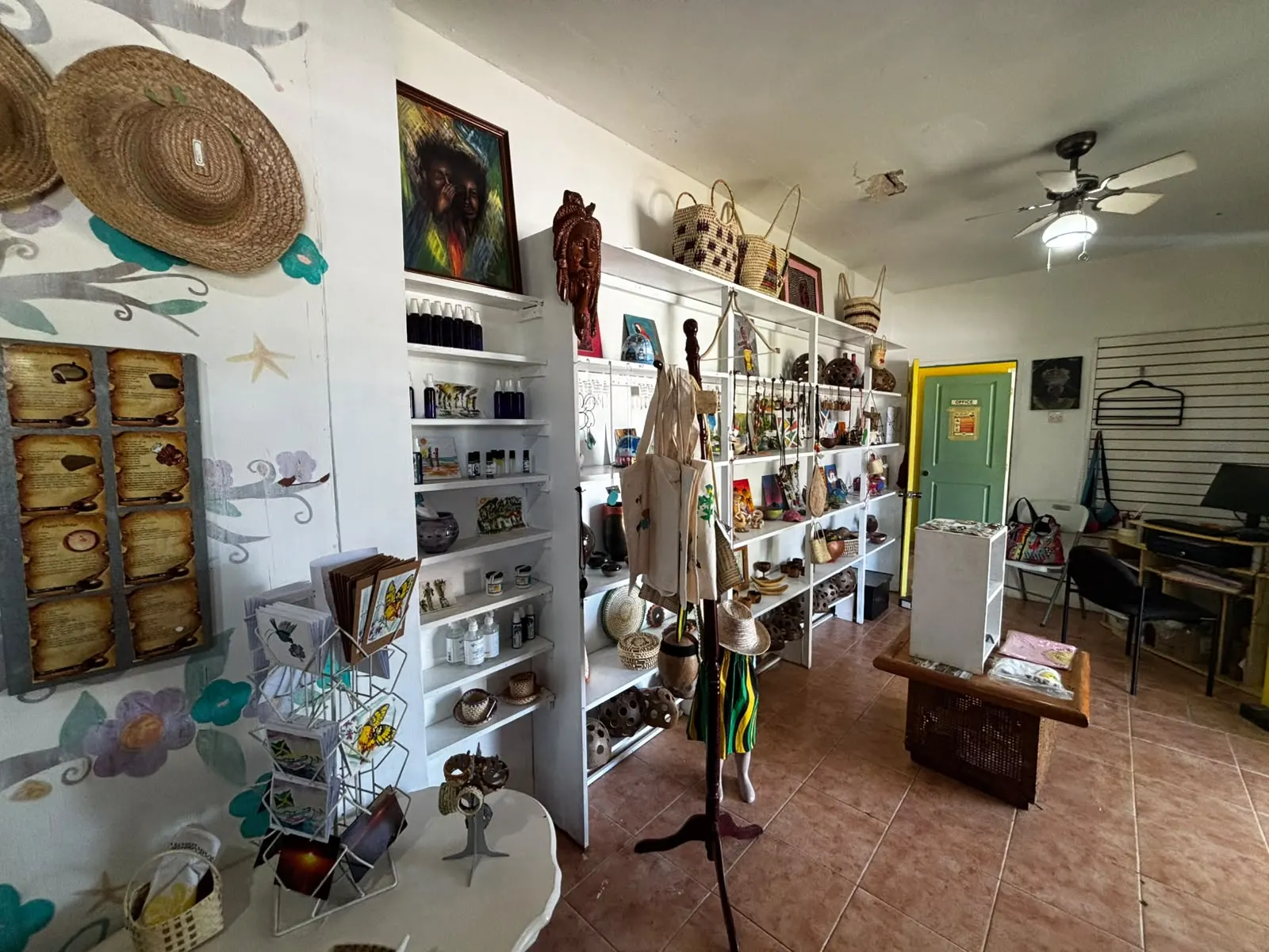 The main room of the shop: white shelves of sprays, oils, pottery, jewellery, paintings and woven bags, a postcard stand, straw hats on the painted wall, and a framed painting above