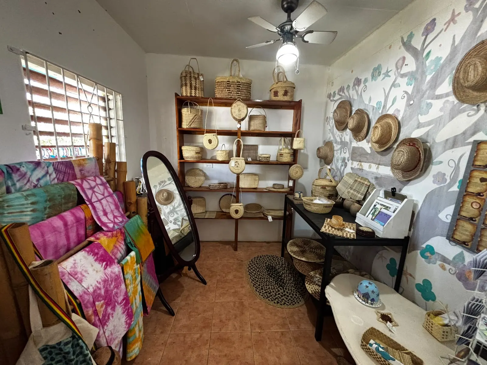 The straw corner of the shop: woven baskets and round bags on a wooden shelf, straw hats on a wall painted with a grey tree and flowers, an oval mirror, and tie-dye cloths hung on bamboo poles