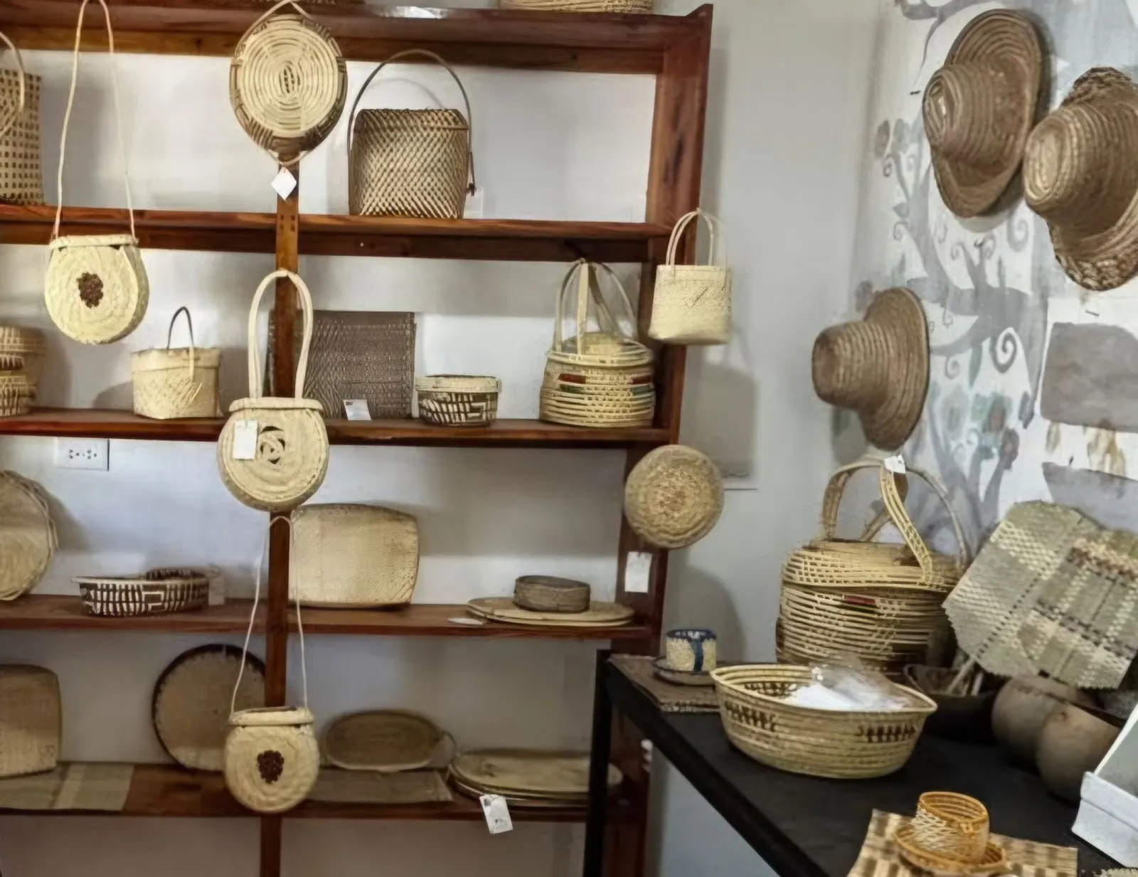 Woven straw goods in the shop: round cross-body bags, baskets with handles, lidded baskets, flat mats and trays on a wooden shelf, straw hats on the wall, and a table of baskets and table mats