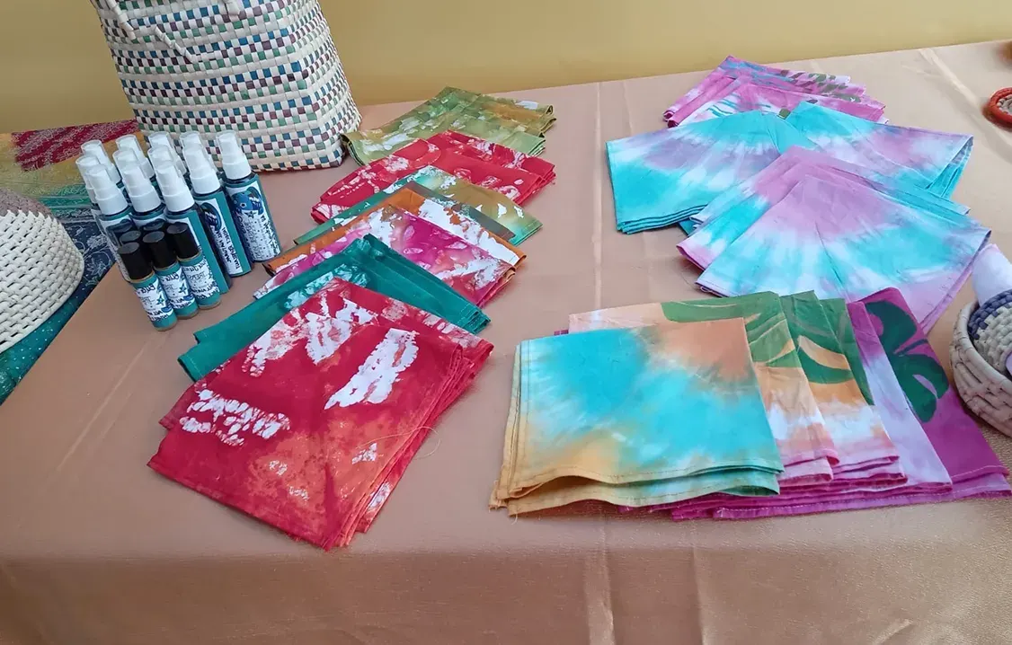 Folded tie-dye and printed cloths in red, teal, pink and orange on a table, with small spray bottles and a woven basket